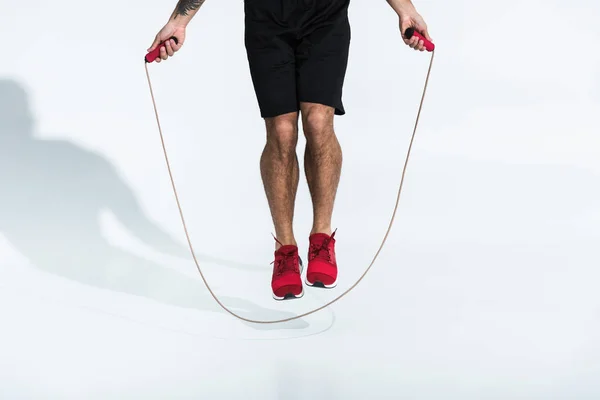 Vista parcial del hombre en pantalones cortos negros y zapatillas rojas saltando con saltar la cuerda en blanco - foto de stock