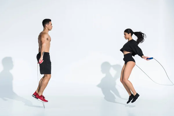 Sportive multicultural man and woman jumping with skipping ropes on white — Stock Photo