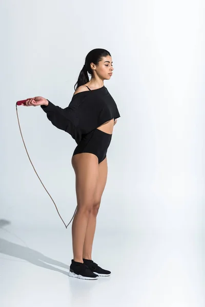 Atractiva mujer afroamericana en ropa deportiva negra y zapatillas de entrenamiento con cuerda de salto en blanco - foto de stock