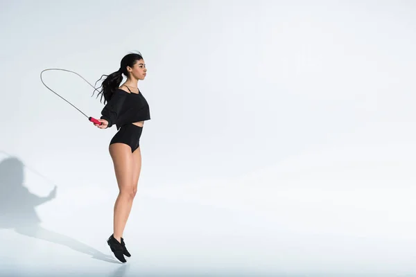 Beautiful sportive african american girl in black sportswear jumping with skipping rope on white — Stock Photo