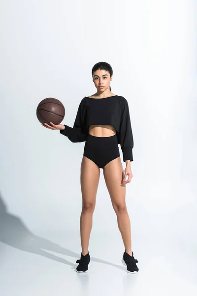 Bella ragazza afro-americana sportiva in possesso di palla marrone e guardando la fotocamera su bianco — Foto stock