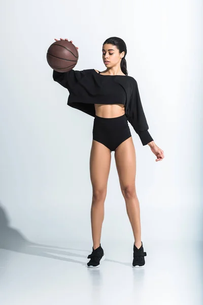 Привлекательная спортивная африканская американка в черной спортивной одежде и кроссовках, играющая в мяч на белом — стоковое фото