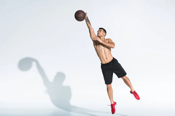 Guapo hombre de raza mixta en pantalones cortos negros y zapatillas rojas saltando con bola en blanco - foto de stock