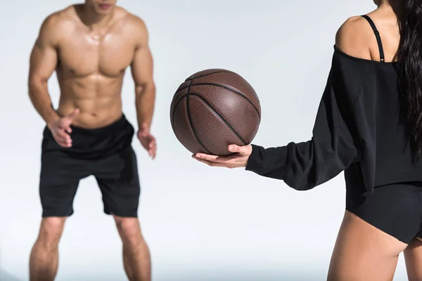 Vista recortada de hombre sin camisa con torso muscular cerca de la chica afroamericana deportiva sosteniendo la pelota en blanco - foto de stock