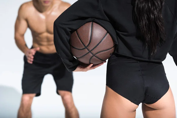Частичный вид спортивной девушки, держащей коричневый мяч возле спортивного мужчины без рубашки на белом — стоковое фото