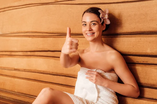 Chica sonriente en toalla con flor en el pelo mostrando el pulgar hacia arriba en la sauna - foto de stock
