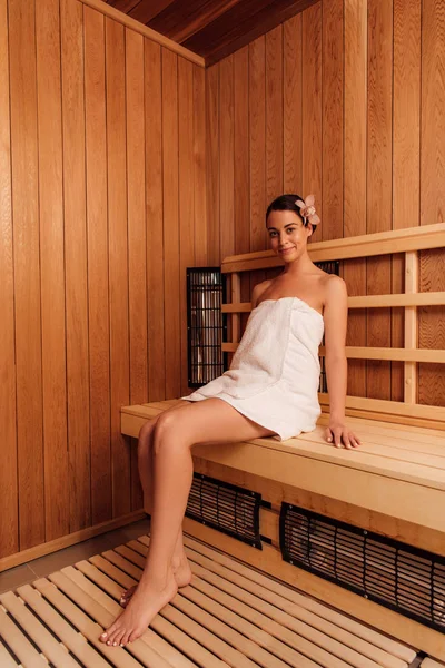 Low angle view of smiling barefoot girl in towel with flower in hair in sauna — Stock Photo