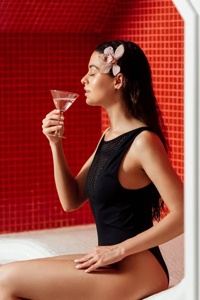 Боковой вид расслабленной молодой женщины с цветком в волосах, пьющей мартини с закрытыми глазами в спа-центре — стоковое фото
