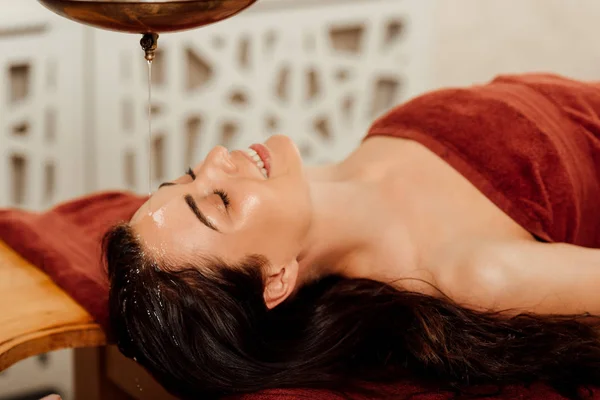 Entspannte junge Frau liegt während ayurvedischer Behandlung unter Shirodhara-Gefäß — Stockfoto