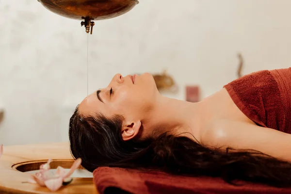 Vista laterale della giovane donna rilassata sdraiata sotto il vaso shirodhara durante la procedura ayurvedica — Foto stock
