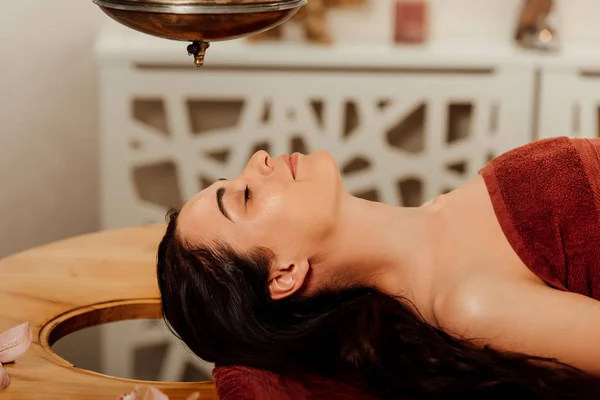 Seitenansicht einer entspannten jungen Frau, die während eines ayurvedischen Eingriffs unter einem Shirodhara-Gefäß liegt — Stockfoto