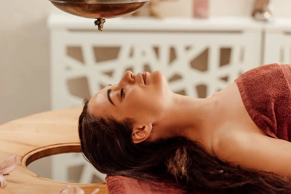 Entspannte junge Frau liegt während ayurvedischer Behandlung unter Shirodhara-Gefäß — Stockfoto