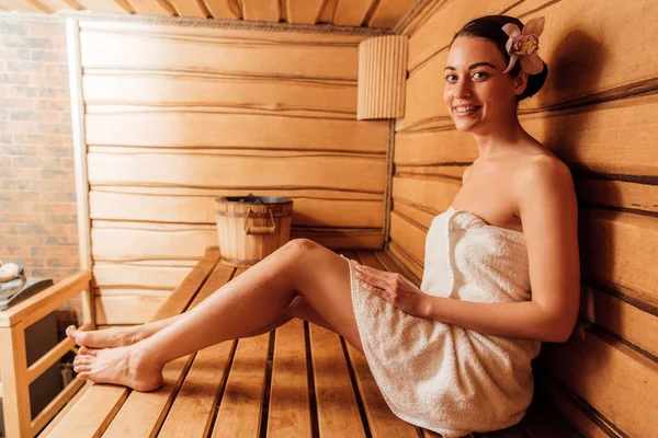 Mujer sonriente en toalla con flor en pelo en sauna - foto de stock