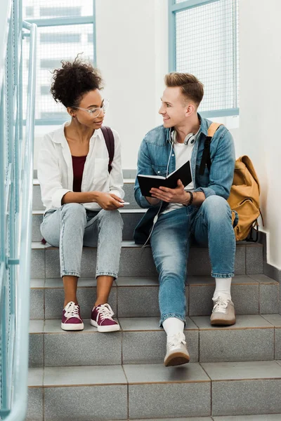 Two multiethinc students with notebook sitting on stairs in university — Stock Photo