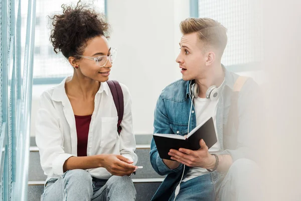 Two multiethinc students with notebook sitting on stairs in university — Stock Photo