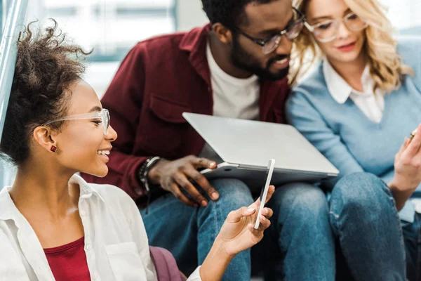 Tres estudiantes multiétnicos sonrientes usando el ordenador portátil y el teléfono inteligente en la universidad - foto de stock