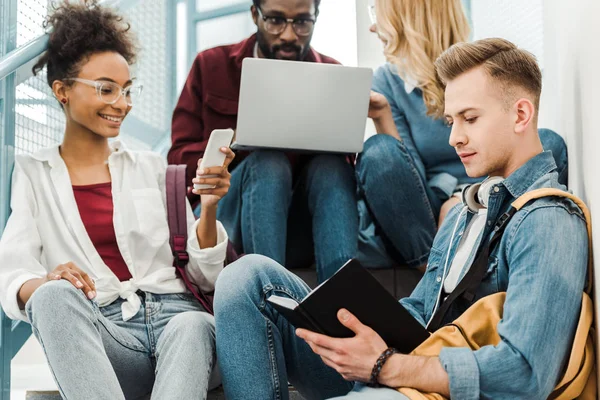 Cuatro estudiantes multiétnicos sonrientes usando el ordenador portátil y el teléfono inteligente en la universidad - foto de stock