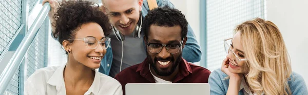 Panoramic shot of smiling multicultural students with laptop in university — Stock Photo