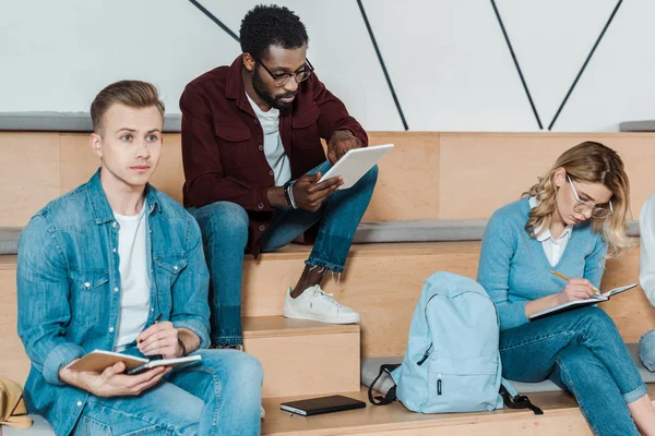 Três estudantes multiétnicos com cadernos e tablet digital estudando em sala de aula — Fotografia de Stock