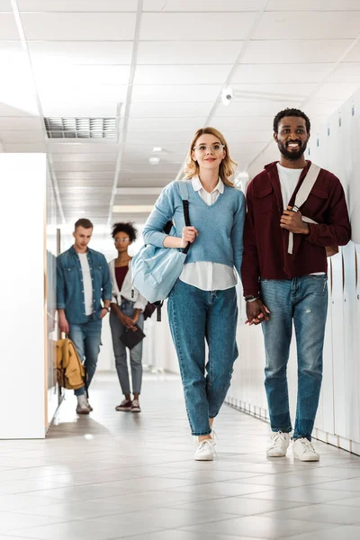 Full length view of smiling multiethnic students holding hands in college — Stock Photo