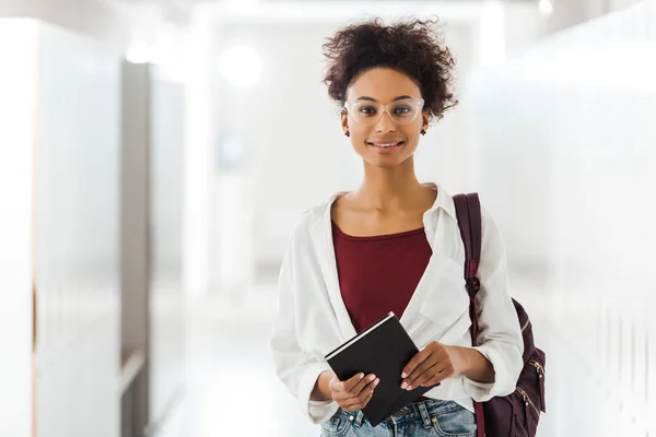 Vista frontal del estudiante afroamericano con cuaderno en pasillo en la universidad - foto de stock