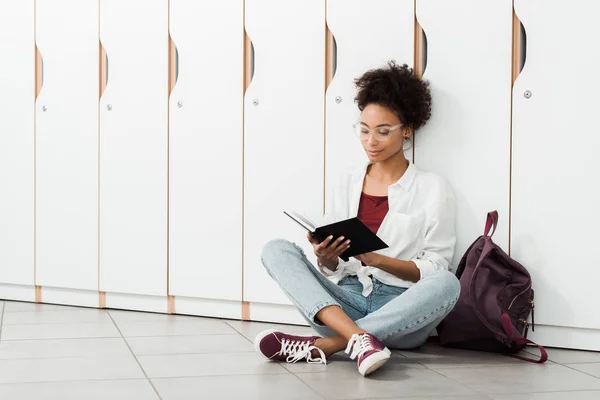 Estudiante afroamericano sentado en el suelo y leyendo un cuaderno en el pasillo - foto de stock