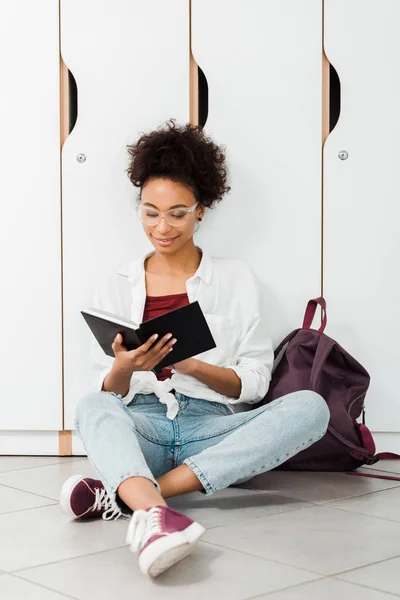 Étudiant afro-américain assis sur le sol et le cahier de lecture dans le couloir — Photo de stock