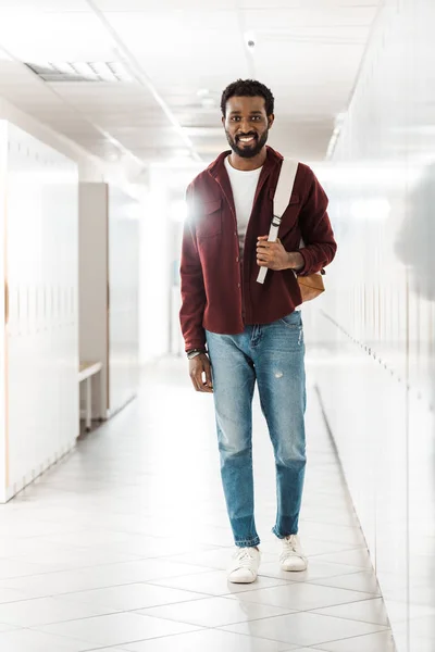 Full length view of african american student in jeans with backpack looking at camera in corridor — Stock Photo