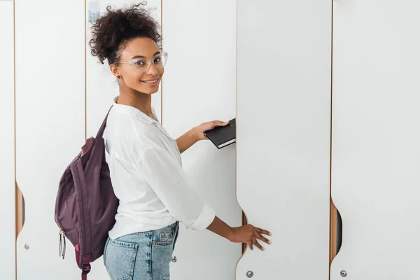 Улыбающийся американский студент из Африки кладет блокнот в шкафчик — стоковое фото