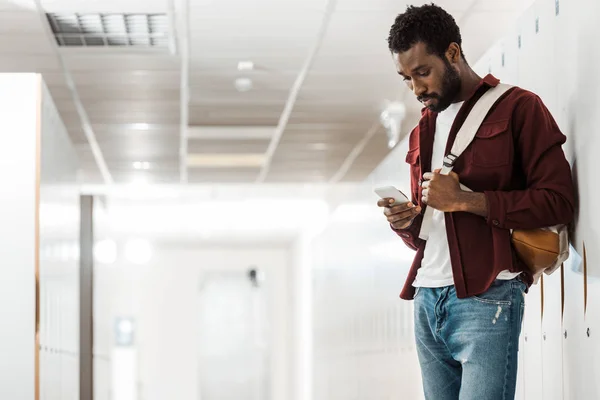 African american student with backpack using smartphone in corridor — Stock Photo