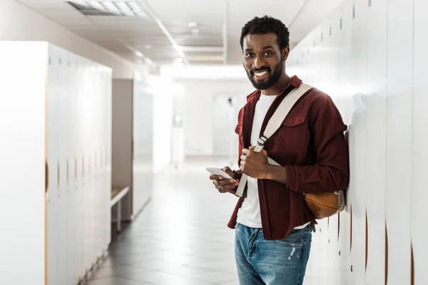 African american student with backpack using smartphone in corridor — Stock Photo