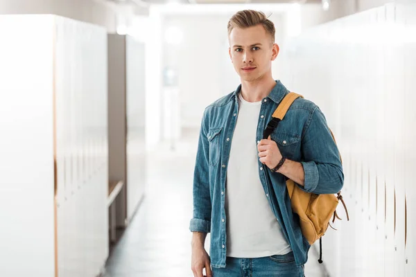 Студент с рюкзаком смотрит на камеру в коридоре университета — стоковое фото