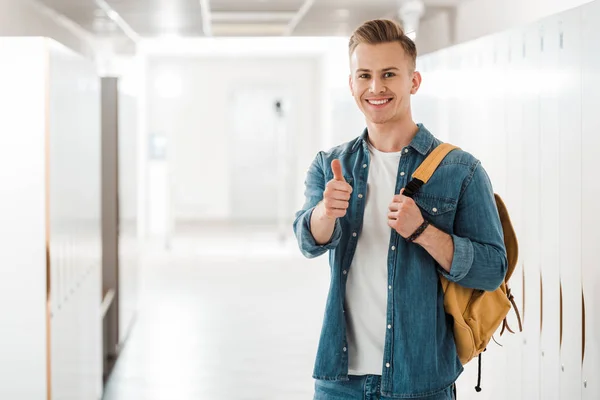 Estudiante con mochila mostrando el pulgar hacia arriba en el pasillo en la universidad - foto de stock