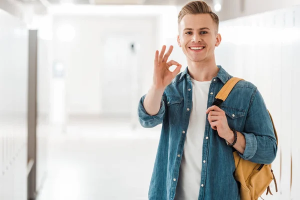 Student with backpack showing okay sign in corridor in university — Stock Photo