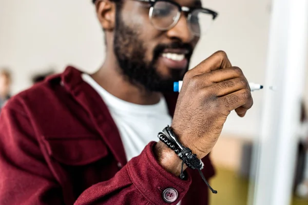 Enfoque selectivo del estudiante afroamericano sonriente en gafas que escriben en el rotafolio - foto de stock