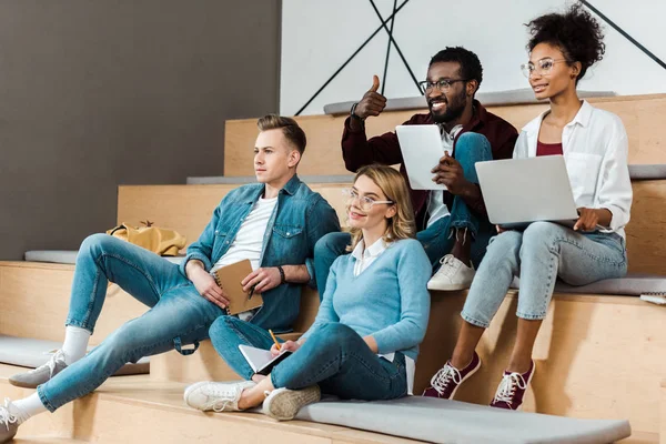 Smiling multicultural students with laptop and digital tablet in lecture hall — Stock Photo