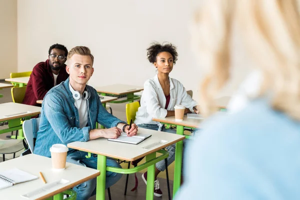 Estudiantes multiétnicos sentados en escritorios y mirando al profesor - foto de stock