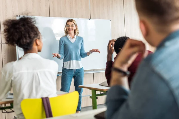 Smiling blonde student in glasses standing near flipchart and looking at friends — Stock Photo