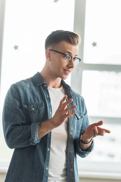 Estudiante emocional en gafas y camisa de mezclilla gestos mientras habla en la universidad - foto de stock