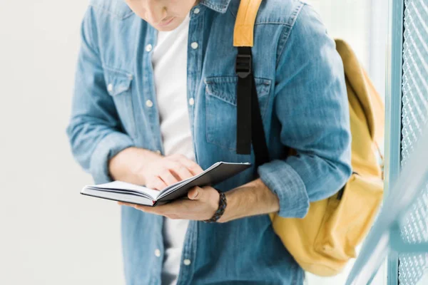 Teilbild eines Studenten im Jeanshemd mit gelbem Rucksack, der Notizbuch liest — Stockfoto