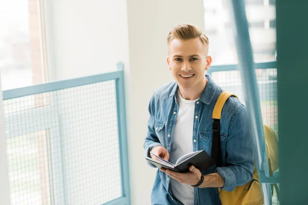 Smiling student in denim shirt with yellow backpack holding notebook in university — Stock Photo