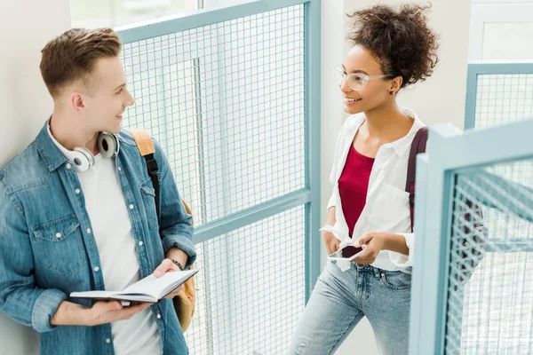 Two multicultural students with notebook and smartphone looking at each other — Stock Photo