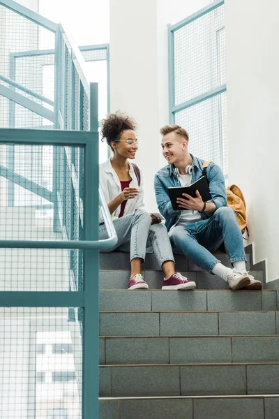 Lächelnde multiethnische Studenten mit Notizbuch auf der Treppe im College — Stockfoto