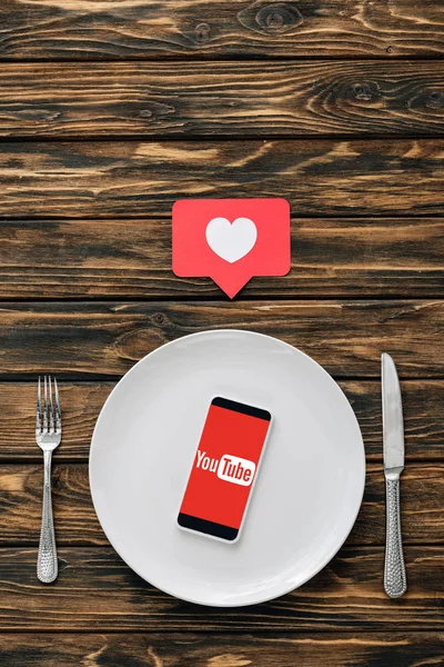 Vista superior del teléfono inteligente con aplicación youtube en la pantalla en la placa blanca cerca de cuchillo, tenedor y tarjeta de corte de papel rojo con símbolo del corazón en la superficie de madera marrón — Stock Photo