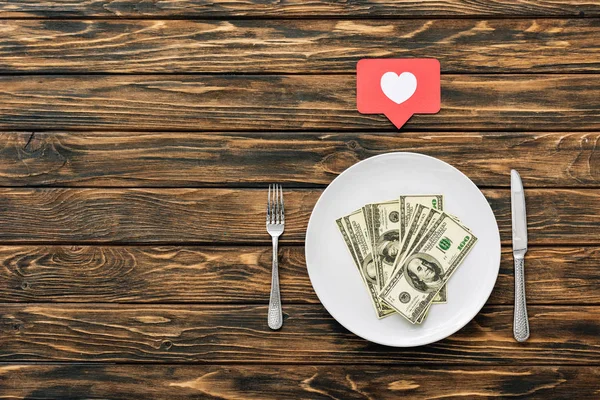Верхний вид банкнот доллара на белой тарелке рядом с красной карточкой разреза бумаги с символом сердца, ножом и вилкой на коричневой деревянной поверхности — стоковое фото