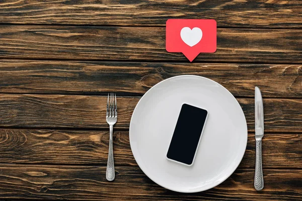 Vue du haut du smartphone avec écran blanc sur plaque blanche près d'un couteau, d'une fourchette et d'un cœur coupé en papier rouge avec symbole du cœur sur une surface en bois marron — Photo de stock