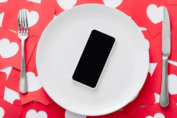 Vue du haut du smartphone avec écran blanc sur plaque blanche près du couteau et fourchette sur papier rouge cartes coupées avec des symboles de coeur — Photo de stock