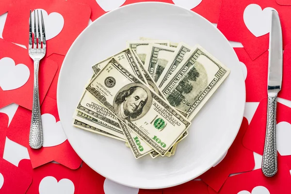 Top view of dollar banknotes on white plate near knife and fork on red paper cut cards with hearts symbols — Stock Photo