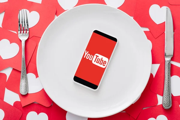 Top view of smartphone with youtube app on screen on white plate near knife and fork on red paper cut cards with hearts symbols — Stock Photo