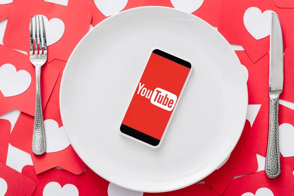 Vista superior del teléfono inteligente con la aplicación de youtube en la pantalla en la placa blanca cerca del cuchillo y tenedor en tarjetas de corte de papel rojo con símbolos de corazones — Stock Photo